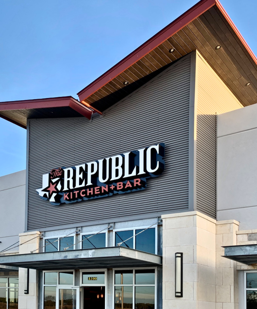 Exterior signage for The Republic Kitchen + Bar mounted on a modern building with metal paneling and stone accents. The bold logo features a red star inside the Texas outline, with "Kitchen + Bar" in red block letters beneath “The Republic.”