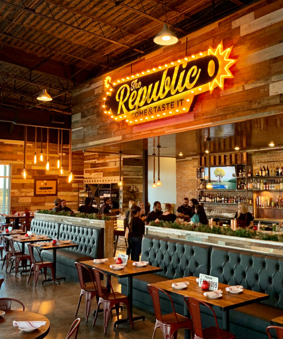 Interior of The Republic Kitchen + Bar featuring rustic wood walls, industrial lighting, and tufted booth seating. A large neon sign above the bar reads “The Republic – Come & Taste It,” adding a bold Texas flair to the lively dining atmosphere.