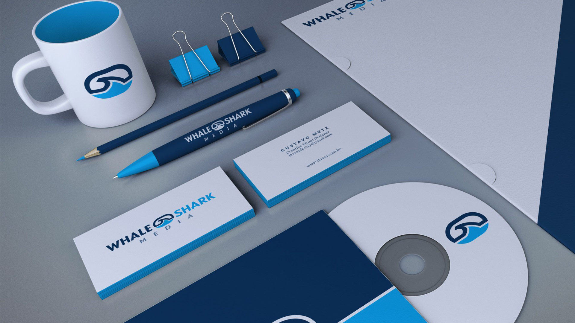 Branded stationery and office items for Whale Shark Media, including a white mug, pens, pencils, business cards, folder, CD, and binder clips, all featuring a blue and white color scheme with the company’s wave and whale-tail inspired logo. The layout presents a cohesive and professional visual identity for the media company.
