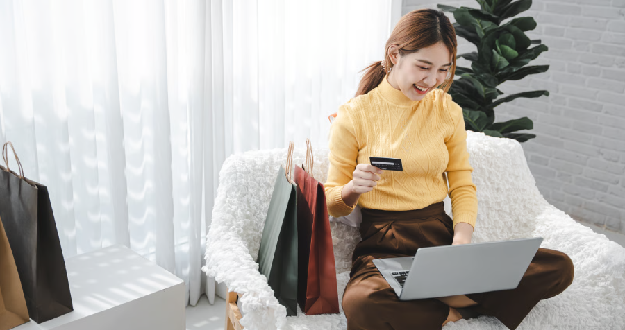 A smiling young woman with auburn hair sits with her legs crossed, laptop on her lap and credit card in her hand.