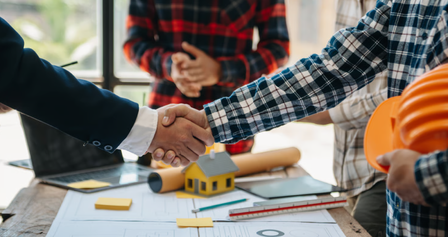 Four individuals, presumably professionals, are gathered around a table reviewing construction plans. A handshake is visible between two people at the forefront of the image.