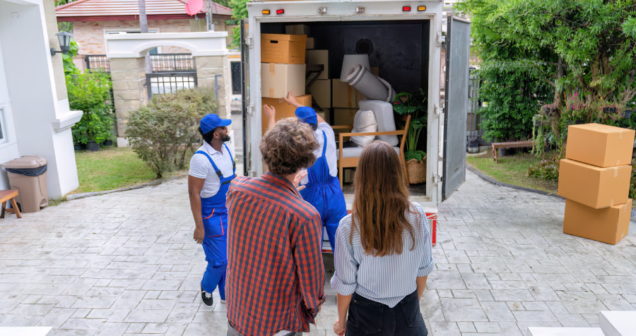 Four people are visible outside of a house with a moving truck. Three people, two men in blue overalls and one man in a red and black plaid shirt, are standing near the moving truck, while a woman in a blue and white striped shirt is standing near the man in the plaid shirt. The men in the blue overalls appear to be moving boxes and furniture into the moving truck.