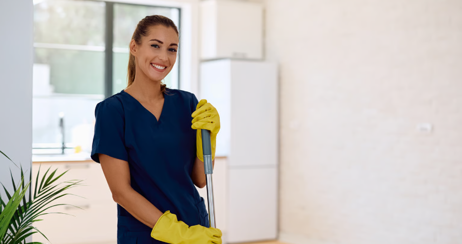 A young woman with tied back hair smiles as she holds a mop in a bright home setting. She is wearing a blue scrub top and yellow gloves.