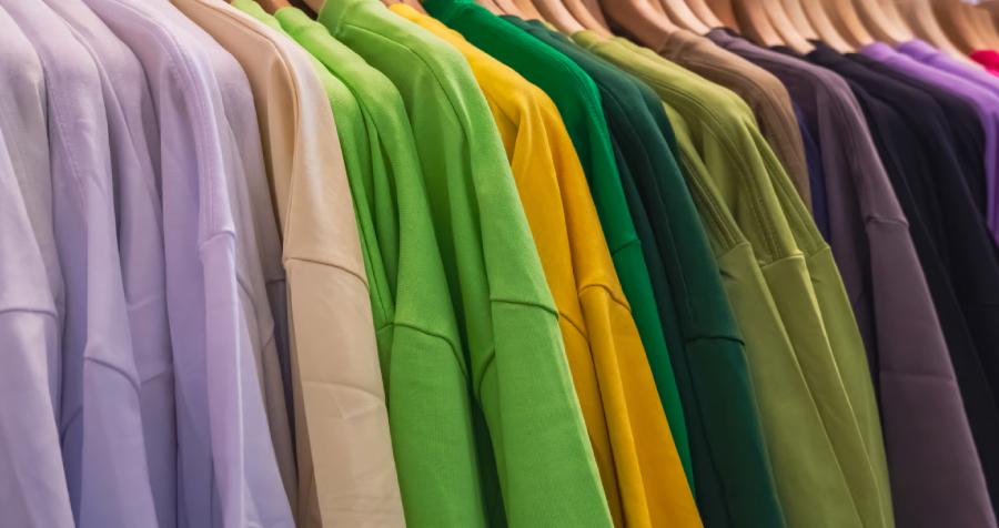 A row of colorful t-shirts hangs on wooden hangers. The shirts are a variety of colors including purple, tan, several shades of green, yellow, and black. The clean, well-lit shot could be used for fashion, retail, or business projects. It could also represent colors, clothing, or choice. The shirts' colors are displayed in a gradient.