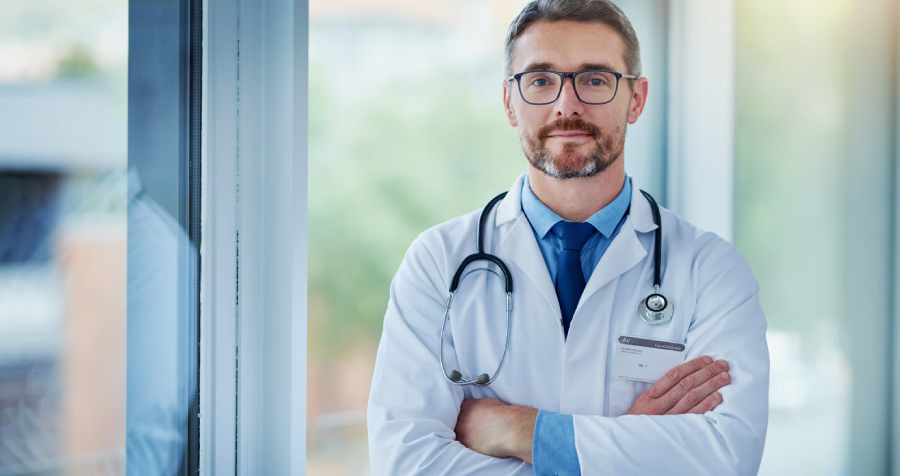 A mature man with short brown hair and glasses poses confidently in front of a window. He wears a white medical coat, a light blue dress shirt, and a blue tie.